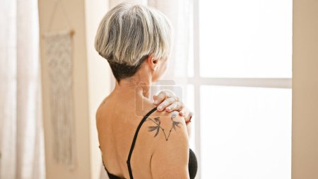 Photo for A mature woman with grey hair and a tattoo looks away in a sunlit bedroom, conveying serenity and introspection. - Royalty Free Image