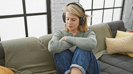 Téléchargez les photos : Jeune femme blonde en vêtements décontractés profitant de la musique avec écouteurs, assise sur un canapé à la maison avec une expression sereine. - en image libre de droit