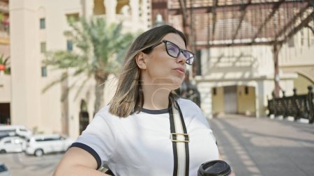 Téléchargez les photos : Une jeune femme réfléchie avec des lunettes se tient au milieu de l'architecture doha moderne, incarnant l'élégance urbaine et les aspirations de voyage. - en image libre de droit