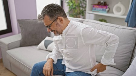 Besorgter, junger hispanischer Mann mit grauen Haaren, der unter starken Rückenschmerzen leidet, sitzt zu Hause auf dem Sofa, Schmerzen spiegeln sich in seiner Brille wider