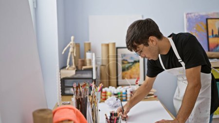 Téléchargez les photos : Jeune homme hispanique attrayant en tablier, concentré dans un atelier d'art, déchaînant dessin créatif avec crayon dans les mains, absorbé dans son passe-temps - en image libre de droit