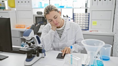 Una mujer científica enfocada en un laboratorio usando un smartphone mientras trabaja junto a un microscopio y tubos de ensayo.