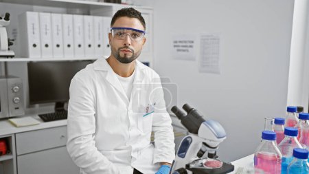 A young man with a beard in a laboratory wearing safety goggles and a lab coat sits before a microscope, exemplifying professionalism and research.