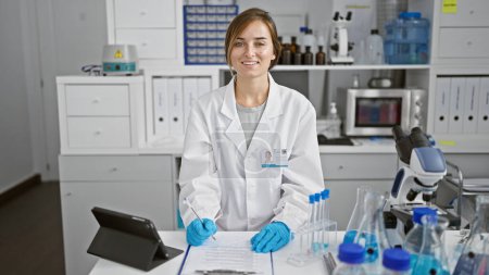 Téléchargez les photos : Radieuse jeune scientifique blonde, occupée à prendre des notes sur son presse-papiers, lance un sourire chaleureux à travers son laboratoire animé, pleinement engagée dans la recherche en biosciences. - en image libre de droit
