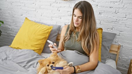 Téléchargez les photos : Jeune femme hispanique se reposant confortablement au lit, magasinant en ligne avec carte de crédit à l'aide d'un smartphone, chien couché à proximité, améliorant sa relaxation matinale dans la chambre - en image libre de droit