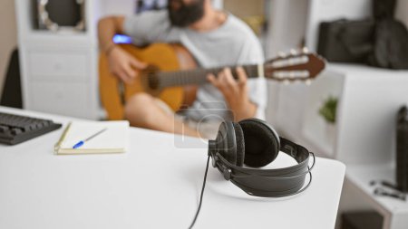 Téléchargez les photos : Un jeune homme hispanique barbu joue de la guitare acoustique dans une pièce intérieure meublée de façon décontractée avec un bureau blanc, un casque et un ordinateur portable. - en image libre de droit
