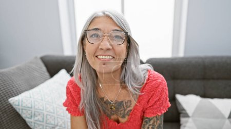Téléchargez les photos : Femme d'âge moyen rayonnante, aux cheveux gris et confiante, souriant joyeusement tout en étant assise confortablement sur le confortable canapé du salon à la maison. - en image libre de droit