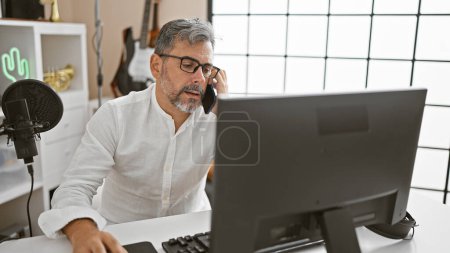 Photo for Gripping on-air conversations, grey-haired, young hispanic radio news reporter in spirited chat on phone within vibrant adult male podcast studio, skillfully juggling smartphone and computer - Royalty Free Image