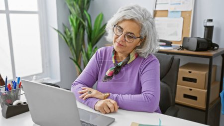 Eine professionelle reife Frau arbeitet aufmerksam an ihrem Laptop in einem modernen Büroumfeld und strahlt Erfahrung und Konzentration aus.