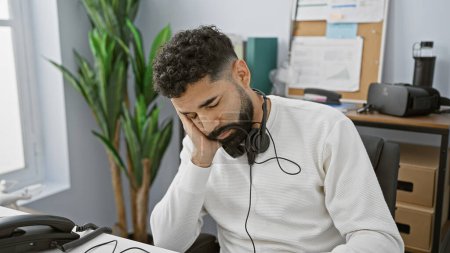 Foto de Hombre hispano cansado con barba usando auriculares en el escritorio de la oficina buscando estresado - Imagen libre de derechos