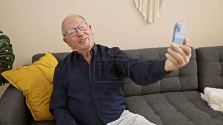 Téléchargez les photos : Joyeux homme âgé assis sur le canapé à la maison, prenant un selfie expressif avec un smartphone, sourire illuminant la pièce - en image libre de droit