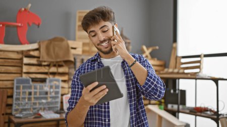 Photo for Handsome young arab man carpenter talking on smartphone, beaming with smiling face, engrossed in woodworking on touchpad indoors at his carpentry workshop studio - Royalty Free Image