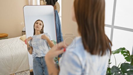 Téléchargez les photos : Une femme brune en tenue décontractée ajuste son t-shirt devant un miroir dans un intérieur de chambre bien éclairé. - en image libre de droit