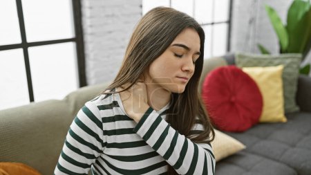 Téléchargez les photos : Jeune femme hispanique avec douleur au cou assise à l'intérieur sur un canapé dans un cadre de salon. - en image libre de droit