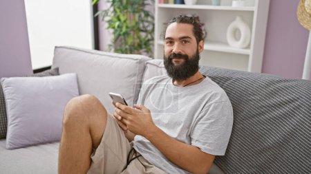 Téléchargez les photos : Homme hispanique confortablement assis sur un canapé, en utilisant un smartphone à l'intérieur d'un salon confortable, rayonnant une ambiance détendue et moderne. - en image libre de droit