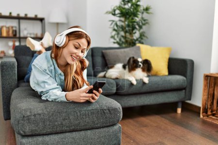 Foto de Joven mujer caucásica escuchando música tumbada en el sofá con el perro en casa - Imagen libre de derechos