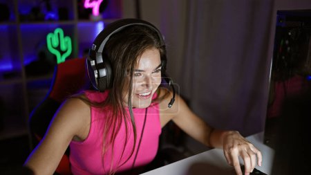 Foto de Sonriente hermosa joven hispana streamer jugando videojuego, lleno de alegría, en vibrante sala de juegos con equipo fresco - Imagen libre de derechos