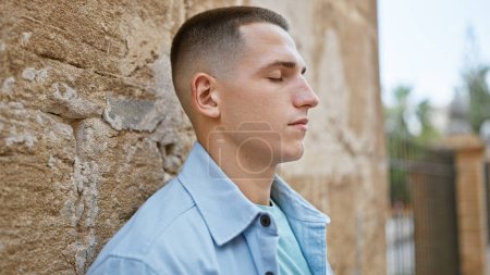 Handsome hispanic man in a serene moment, leaning against an old city wall outdoors.
