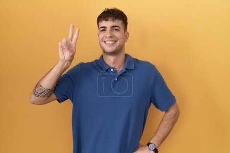 Foto de Joven hombre hispano de pie sobre fondo amarillo sonriendo mirando a la cámara mostrando los dedos haciendo señal de victoria. número dos. - Imagen libre de derechos