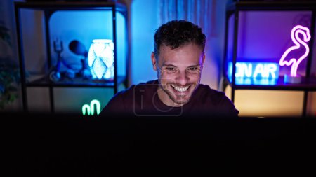 Photo for A smiling hispanic man with a beard enjoys his time in a vibrant gaming room at night. - Royalty Free Image
