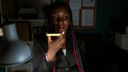 Foto de Elegante jefa afroamericana trabajando hasta altas horas de la noche, enviando un mensaje de voz profundo y serio desde su teléfono inteligente en la mesa de la oficina. - Imagen libre de derechos