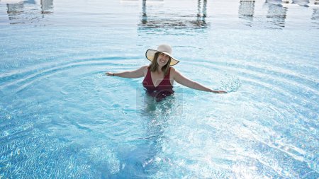 Téléchargez les photos : Femme souriante en maillot de bain profitant du soleil à la piscine de luxe avec les bras tendus - en image libre de droit