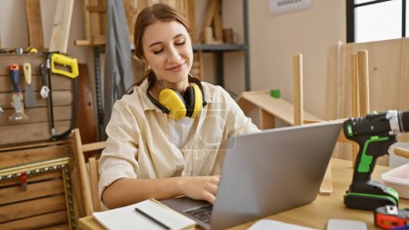 Téléchargez les photos : Femme brune travaillant sur ordinateur portable dans un atelier de menuiserie avec écouteurs et outils autour. - en image libre de droit
