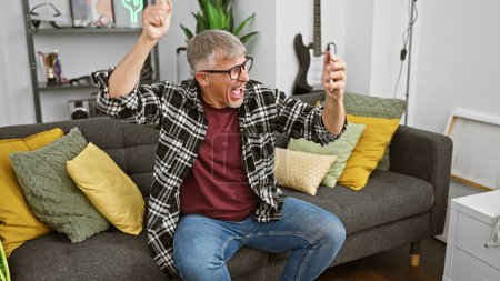 Foto de Emocionado hombre de mediana edad celebrando la victoria en casa, sentado en un sofá gris en una luminosa sala de estar. - Imagen libre de derechos