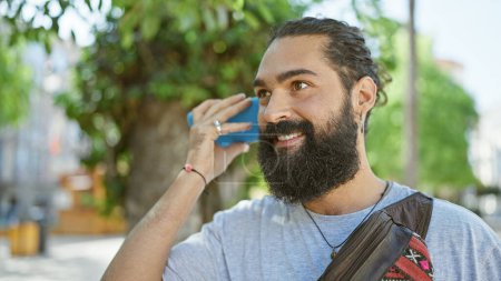 Foto de Guapo joven hispano con barba sonriendo mientras escucha un mensaje de voz en su teléfono al aire libre en la ciudad. - Imagen libre de derechos