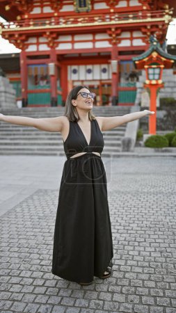 Joyful hispanic woman in glasses, embracing freedom with open arms, smiling and looking around fushimi inari taisha shrine in kyoto, japan