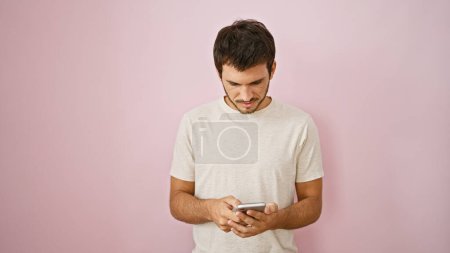 Foto de Hombre hispano guapo con barba, vistiendo una camiseta casual, enviando mensajes de texto en su teléfono contra una pared rosa vívida - Imagen libre de derechos