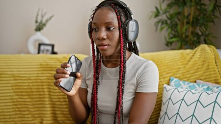 Photo for Beautiful african american woman, seriously focused, enjoys her afternoon singing along to her favorite song. relaxed on her living room sofa, she's engrossed in the music, headphones on. - Royalty Free Image