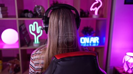 Foto de Vista trasera de una mujer rubia con auriculares en una vibrante sala de juegos por la noche. - Imagen libre de derechos