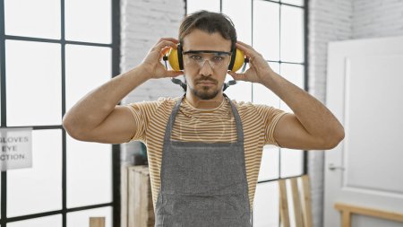 Photo for Young hispanic man wearing safety goggles and headphones in a bright, modern carpentry studio. - Royalty Free Image