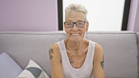 Photo for Cheerful grey-haired senior woman finds joy, comfortably settled on her cozy living room sofa. her confident smile reveals pure happiness in the relaxed, indoor atmosphere of her home. - Royalty Free Image