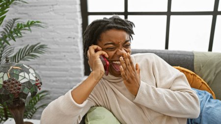 Téléchargez les photos : Femme afro-américaine avec dreadlocks rire énergiquement tout en parlant sur un téléphone rouge, à l'intérieur sur un canapé. - en image libre de droit