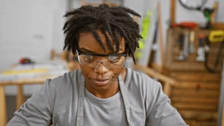 Téléchargez les photos : Une jeune femme concentrée avec des dreadlocks portant des lunettes de sécurité dans un atelier de menuiserie - en image libre de droit