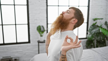Foto de Hombre pelirrojo guapo encontrando su zen de la mañana, concentrándose en ejercicios de yoga mientras está sentado en su cama en la calma de su dormitorio - Imagen libre de derechos
