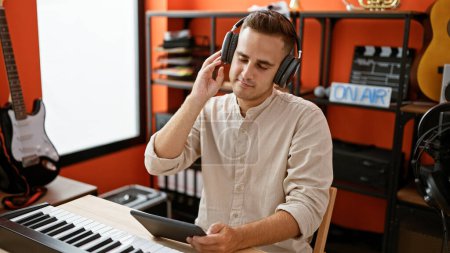 Téléchargez les photos : Beau homme appréciant la musique avec écouteurs, en utilisant une tablette dans un studio de musique avec des instruments. - en image libre de droit