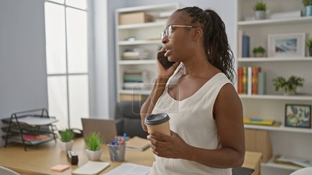 Téléchargez les photos : Femme afro-américaine aux cheveux bouclés au téléphone tenant un café au bureau - en image libre de droit