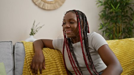 Téléchargez les photos : Femme afro-américaine confiante se détendre sur un canapé confortable, réconfortant la pièce avec son expression vibrante, parlant joyeusement du cœur tout en étant assis à la maison - en image libre de droit
