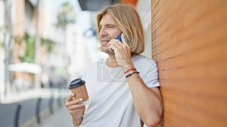 Foto de Joven guapo con el pelo largo y rubio hablando en un celular y sosteniendo una taza de café en una calle de la ciudad. - Imagen libre de derechos