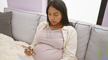 Foto de Mujer hispana joven y relajada, alegremente envuelta en una manta, sentada en el sofá de la sala de estar de su casa, revela sus noticias de embarazo en línea usando su teléfono inteligente, completamente absorta en la felicidad de la maternidad. - Imagen libre de derechos
