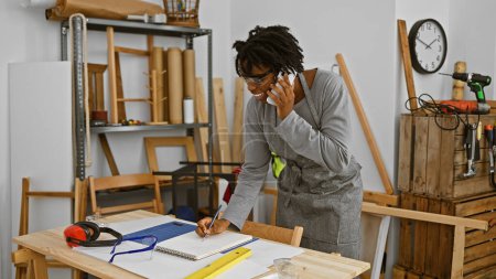 Téléchargez les photos : Une jeune femme avec dreadlocks parle au téléphone tout en prenant des notes dans un atelier de menuiserie - en image libre de droit