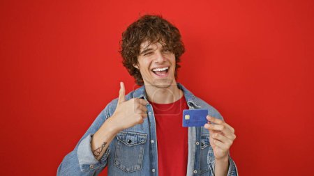 Photo for A cheerful young man with a beard giving thumbs up and holding a credit card against a red wall. - Royalty Free Image