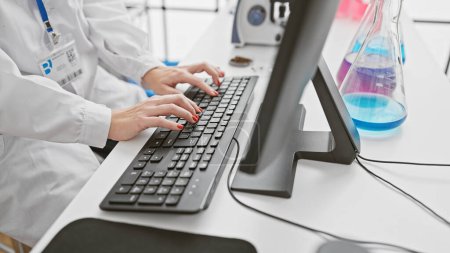 Foto de Científica profesional escribiendo en un teclado en un entorno de laboratorio moderno con coloridos frascos erlenmeyer en el fondo. - Imagen libre de derechos