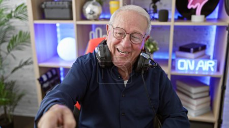 Téléchargez les photos : Streerer homme âgé joyeux, rayonnant avec un grand sourire, engagé dans son cyber jeu. portant un casque, des lunettes et pointant du doigt l'ordinateur dans une salle de jeux animée - en image libre de droit