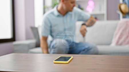 Un homme portant une chemise bleue vérifie sa montre dans un salon moderne avec un smartphone sur la table.