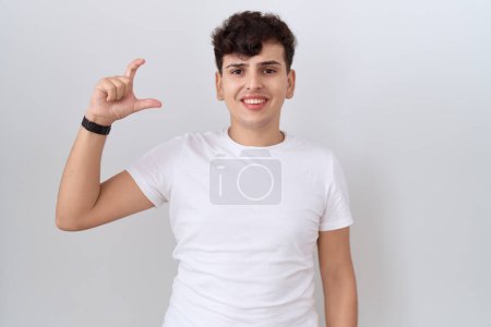 Téléchargez les photos : Jeune homme non binaire portant t-shirt blanc décontracté geste souriant et confiant avec la main faisant signe de petite taille avec les doigts à la recherche et la caméra. concept de mesure. - en image libre de droit