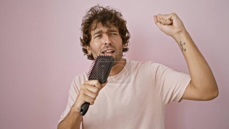 Foto de Hombre hispano cantando con un cepillo de pelo sobre un fondo rosa aislado, exudando un ambiente despreocupado y alegre. - Imagen libre de derechos